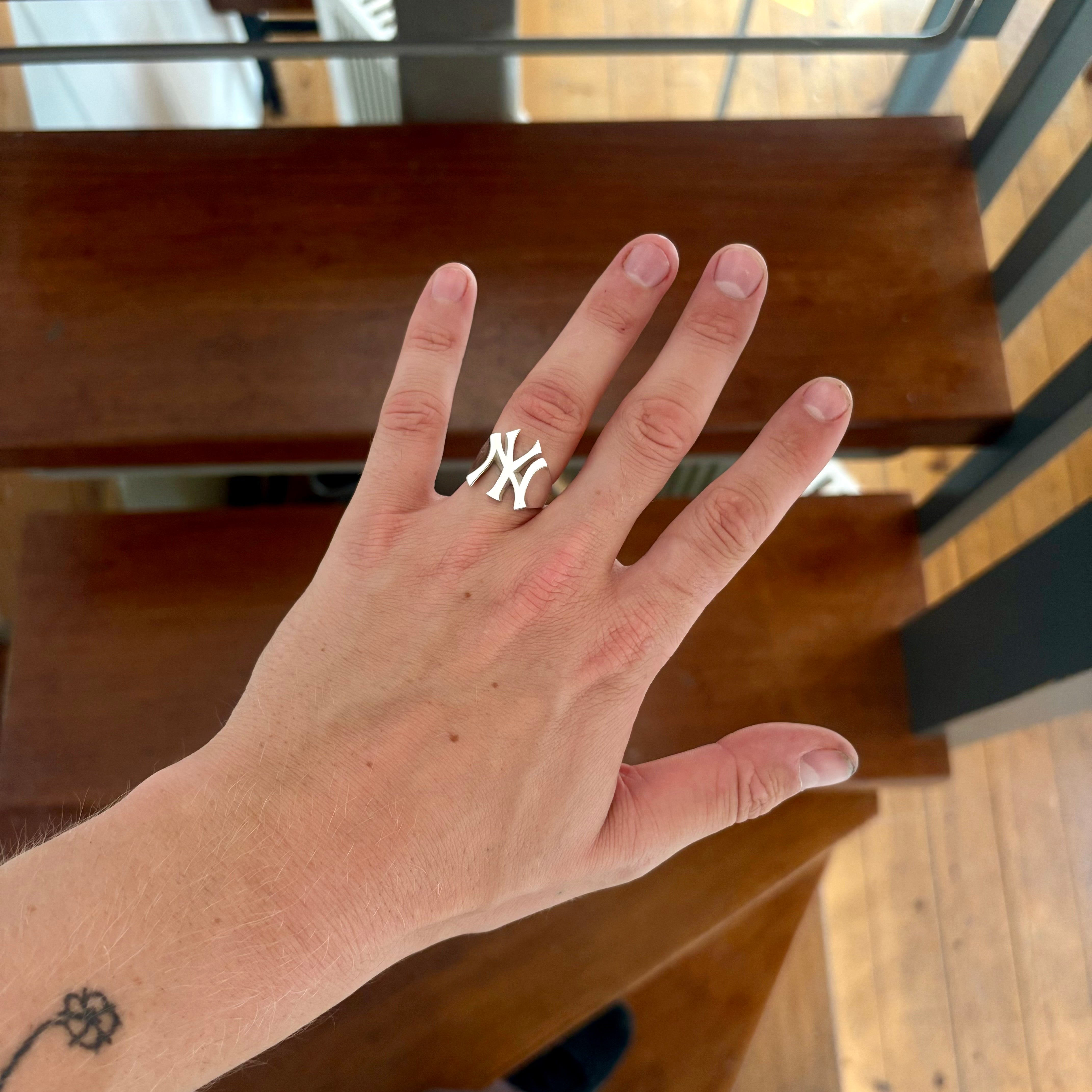 A silver NY Yankees logo ring worn on a person's ring finger, shown in a lifestyle setting with wooden stairs in the background.